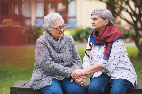 Two senior women sitting together on a bench, holding hands and supporting each other